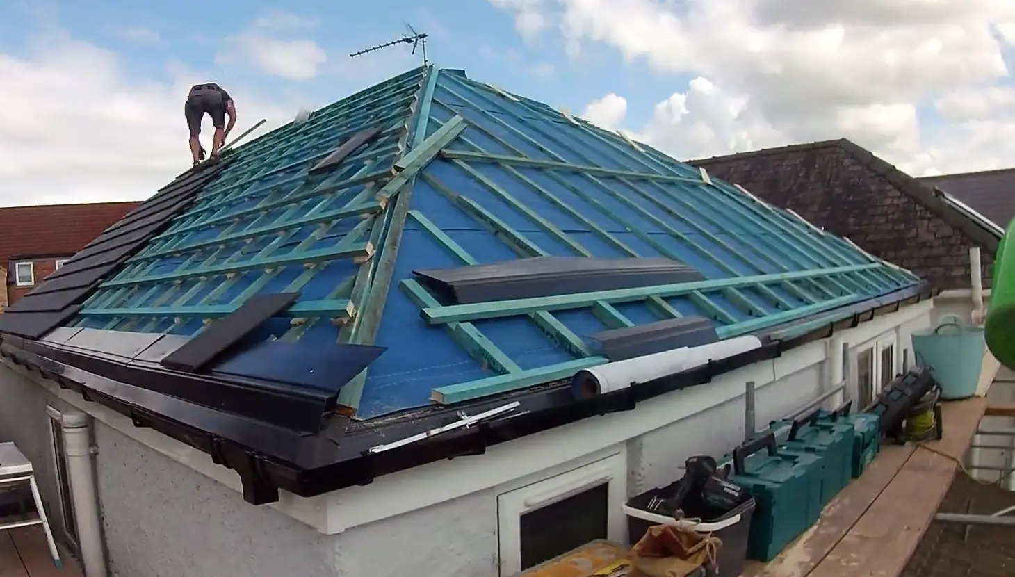 Roofer laying slate tiles over blue waterproof underlayment membrane and wooden battens on a hip roof structure.